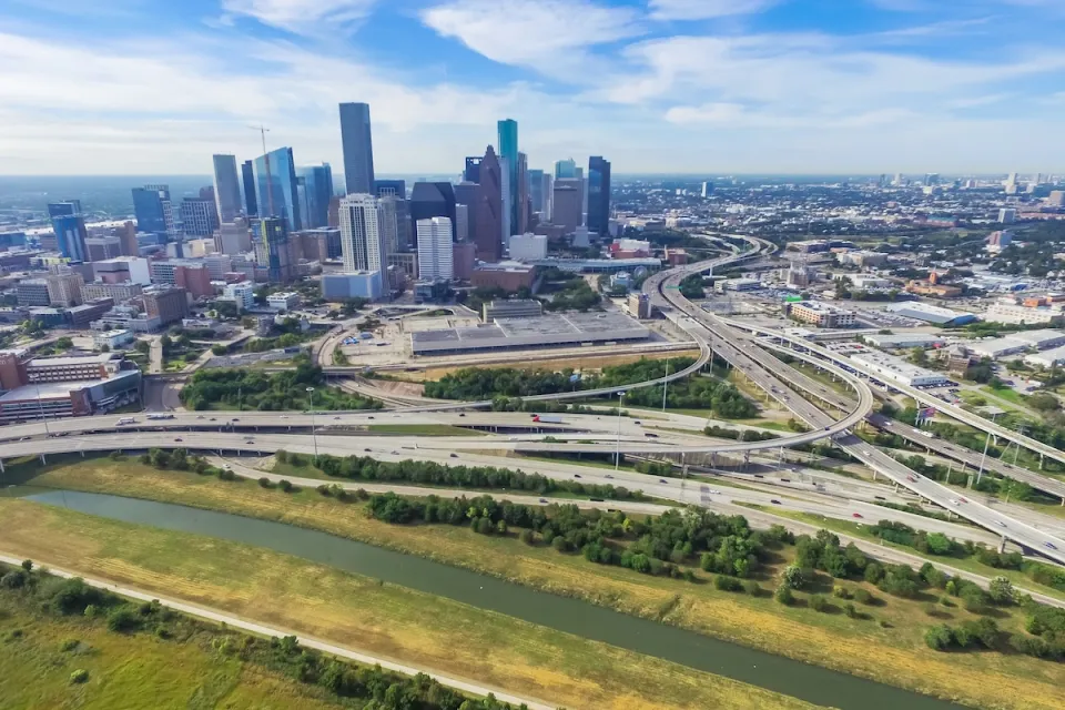 Aerial view of massive freeway interchange with downtown Houston skyline in background.